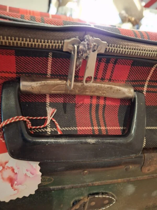 Nærbillede af håndtaget og den kraftige lynlås på en rødrutet vintage kuffert, med et lille hjertevedhæng på lynlåsen.