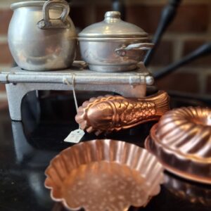 Et gråt metal børnekomfur med kogeplade og to matchende gryder, samt en kobberfiskefrom og en rund kobberbageform i forgrunden.