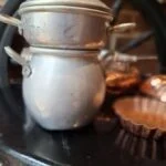 En vintage sølvfarvet metalgryde og en mindre gryde ovenpå, begge med låg og hanke, sandsynligvis til børnekomfur.