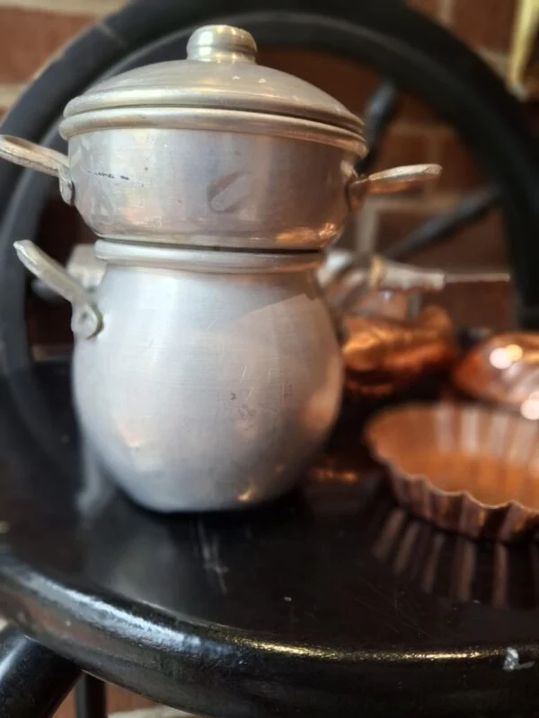 En vintage sølvfarvet metalgryde og en mindre gryde ovenpå, begge med låg og hanke, sandsynligvis til børnekomfur.