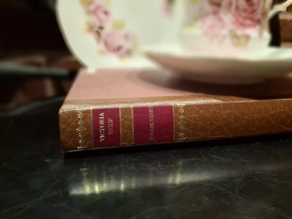 Bogryggen af Victoria Holt's 'Judaskysset' med brun indbinding og røde titelfelter, ved siden af et hvidt tesæt med roser.