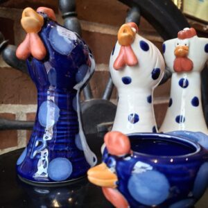 Fire sjove keramikfigurer formet som høns, to blåstribede og to hvide med blå prikker, stående på en mørk overflade.