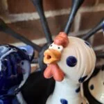 Nærbillede af en hvid keramik hønefigur med blå prikker og orange kam, næb og hagel, stående mod en murstensvæg. Figuren har et åbent hoved som kan bruges til en æggebæger.