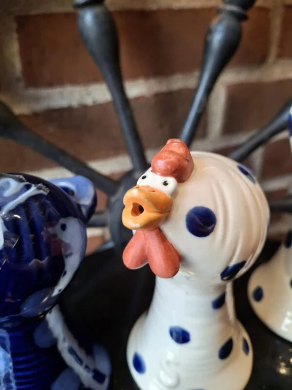 Nærbillede af en hvid keramik hønefigur med blå prikker og orange kam, næb og hagel, stående mod en murstensvæg. Figuren har et åbent hoved som kan bruges til en æggebæger.