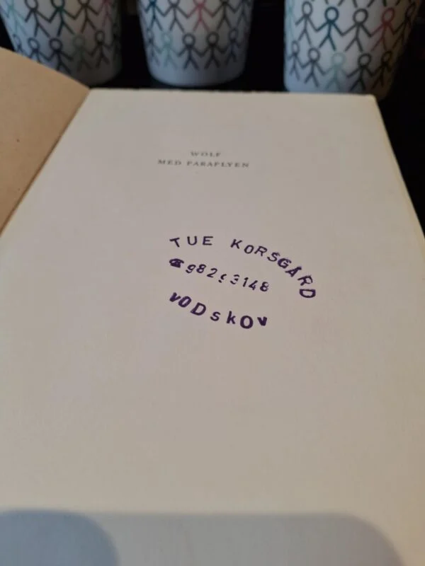 Inderside af bogen 'Wolf med Paraplyen' med et lilla stempel, der viser 'Tue Korsgård 98253148 Vodskov'.
