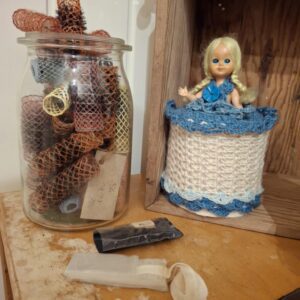 En samling vintage hårfarver og farverige curlere i en stor glasbeholder på et træbord, ved siden af en dukke.