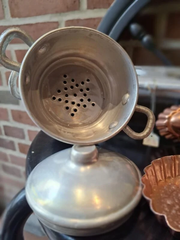 Nærbillede af en vintage sølvfarvet metalgryde til børnekomfur. Overdelen er en perforeret indsats der matcher det grå stel, den står på.
