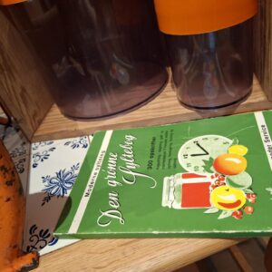 Retro kaffefilter, Margrethe-skål og opbevaringsbøtter alle i orange, sammen med en grøn syltebog, placeret på en træhylde.