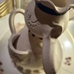 En keramisk figur-vase formet som en dame, set fra oven med blå sløjfe om halsen, med en hvid porcelænstekande.