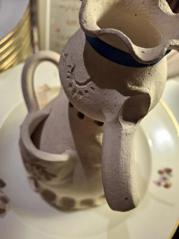 En keramisk figur-vase formet som en dame, set fra oven med blå sløjfe om halsen, med en hvid porcelænstekande.