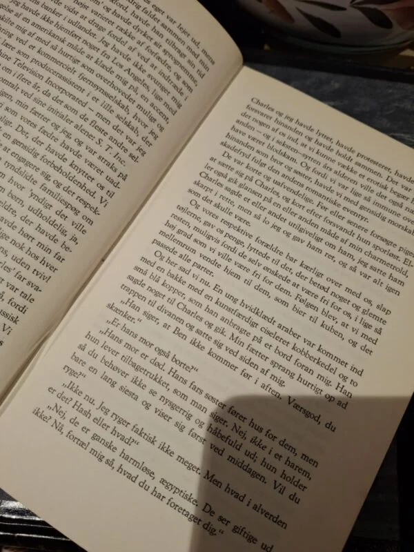 Åben bog med tekst på begge sider. Del af en teservice med kop og underkop ses i baggrunden.
