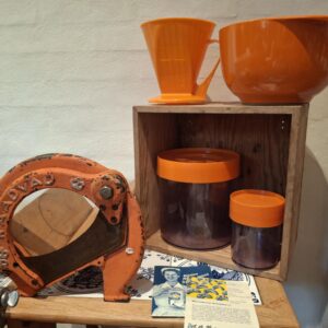 Et orange retro kaffefilter i plastik, anbragt på en træhylde.