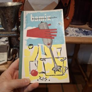 En hånd holder et vintage Bamix hæfte med blå, rød og gul tegning af en stavblender og madvarer.