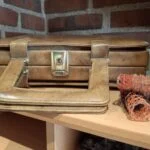 Nærbillede af den gyldne lukkemekanisme på en lukket brun retro toilettaske, der står på en træhylde.