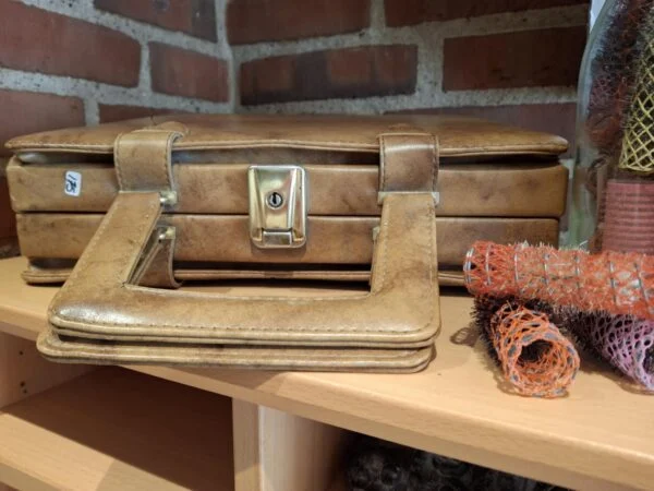 Nærbillede af den gyldne lukkemekanisme på en lukket brun retro toilettaske, der står på en træhylde.