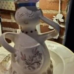 En keramisk figur-vase udformet som en kvinde iført forklæde, der holder en kande. Vasen er detaljeret og farvet med naturmotiver.