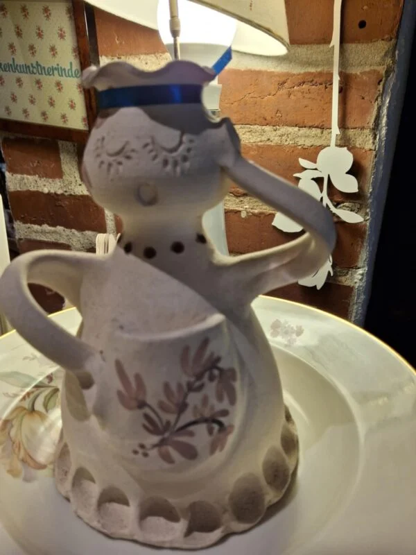 En keramisk figur-vase udformet som en kvinde iført forklæde, der holder en kande. Vasen er detaljeret og farvet med naturmotiver.
