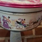 En lille rund vintage Quality Street dåse med rødt låg og detaljerede illustrationer, set fra siden.