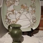 Vintage turkisgrønt kunstfad på sort stativ foran en murstensvæg, med motiv af kvinde og fugl. En grøn keramikvase står foran.