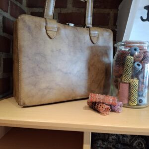 En brun, retro toilettaske, der står lukket på en træhylde foran en murstensvæg, med en krukke af hårcurlere ved siden af.