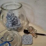 Et glas med håndhæklede grå make-up fjernerpuder, en makeupbørste, broche og glaslåg med blå sløjfe.