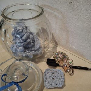 Et glas med håndhæklede grå make-up fjernerpuder, en makeupbørste, broche og glaslåg med blå sløjfe.