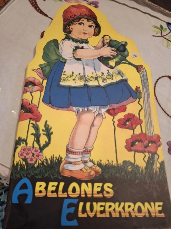 Forsiden af eventyrbogen 'Abelones Elverkrone', udskåret i form af en pige, der vander blomster. Illustreret i vintage stil.