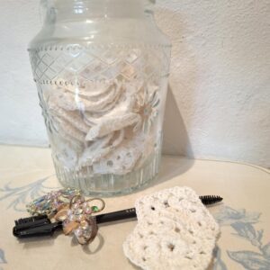 En glasbeholder fyldt med håndhæklede beige make-up fjernerpuder, en makeupbørste og broche på dækket bord.