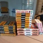En stor stak 'Hvem Hvad Hvor' årbøger i forskellige årgange fra sort, hvid og orange rygdesign på et træbord.
