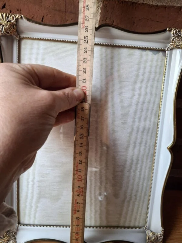 En elegant hvid vintage fotoramme med gyldne, ornamenterede detaljer i hjørnerne, stående oprejst med murstensvæg i baggrund.