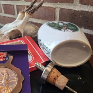 Bagside af den hvide porcelænsbrændevinsflaske med et svagt aftryk, en skænkeprop med kork, samt et hjortekranium og murstensvæg i baggrunden.
