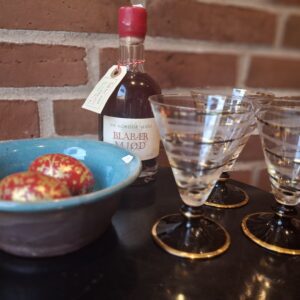 En flaske 'Blåbær Mjød', fire vintage cocktailglas med gyldne kanter, en blå keramikskål og to røde trææg med guldglimmer.