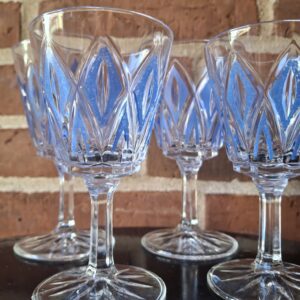 Fire klare aperitifglas med blå, rombeformet mønster, stående på en mørk overflade foran en murstensvæg.