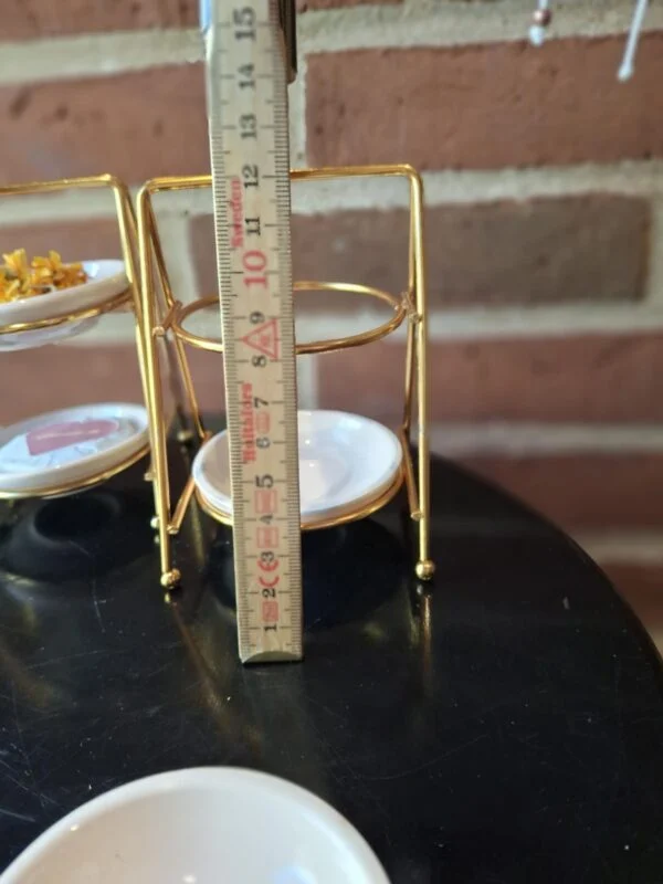 Et to-etagers smykkestativ i gyldent metal og hvide skåle måles med et træmålebånd, der viser en højde på ca. 12 cm.