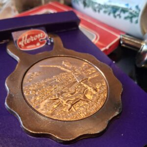 Nærbillede af den bronzefarvede Heron oplukker med et detaljeret motiv af en jæger og hund i et landskab, præsenteret på en lilla baggrund.