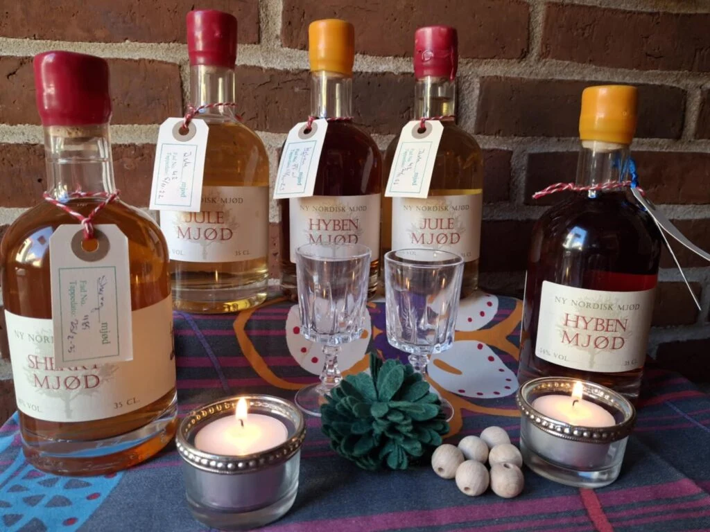 Fem flasker nordisk mjød (Sherry, Jule, Hyben) med to krystal dessertglas, tændte fyrfadslys og grøn filtblomst på bordet.