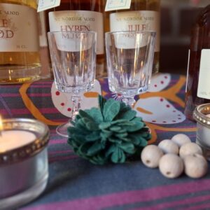 To små krystal dessertglas placeret på en farverig dug, omgivet af tændte fyrfadslys og flasker med mjød i baggrunden.