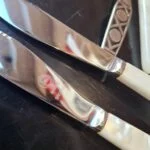 To knive med lyse greb og forskellige mærkninger på bladene; en gaffel ses delvist ved siden af.