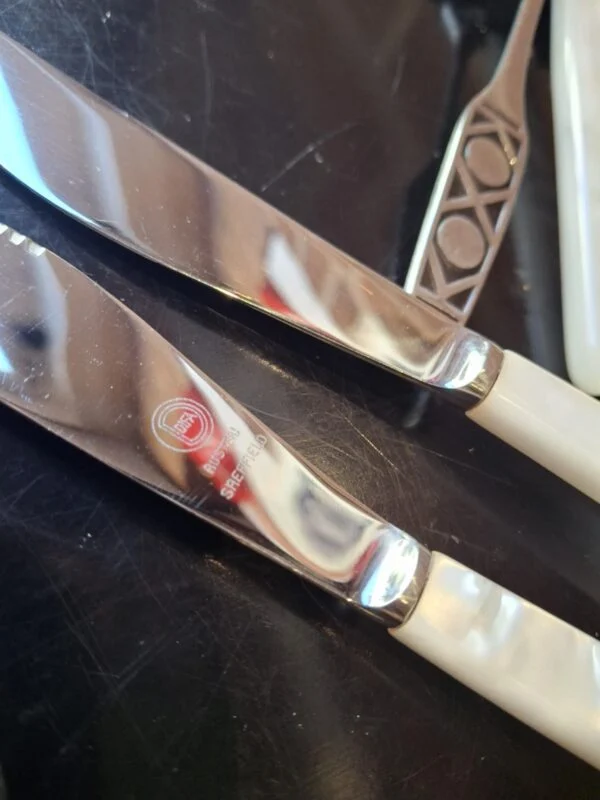 To knive med lyse greb og forskellige mærkninger på bladene; en gaffel ses delvist ved siden af.