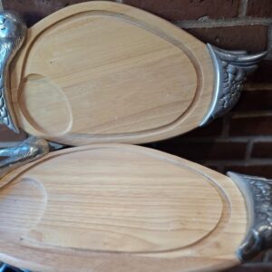 To ovale serveringsbrætter af lyst træ med udskårne riller og metalender formet som ænder, set fra oven på en mørk baggrund.