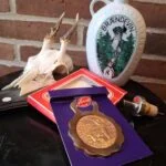 Hvid porcelænsbrændevinsflaske med grønt jagtmotiv og 'BRÆNDEVIN', en bronzefarvet Heron oplukker på lilla baggrund, og en skænkeprop med kork. Hjortekranium og murstensvæg i baggrunden.