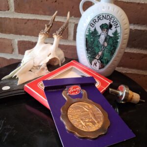 Hvid porcelænsbrændevinsflaske med grønt jagtmotiv og 'BRÆNDEVIN', en bronzefarvet Heron oplukker på lilla baggrund, og en skænkeprop med kork. Hjortekranium og murstensvæg i baggrunden.