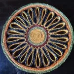 Rund håndflettet bordskåner i naturflet, med et mønster af spiraler og løkker i gule og blå nuancer, tekst 'bastskåner side 2'.