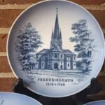 Blå-hvid jubilæumsplatte med Frederikshavn Kirke og årstallene 1818–1968, stillet på et stativ.