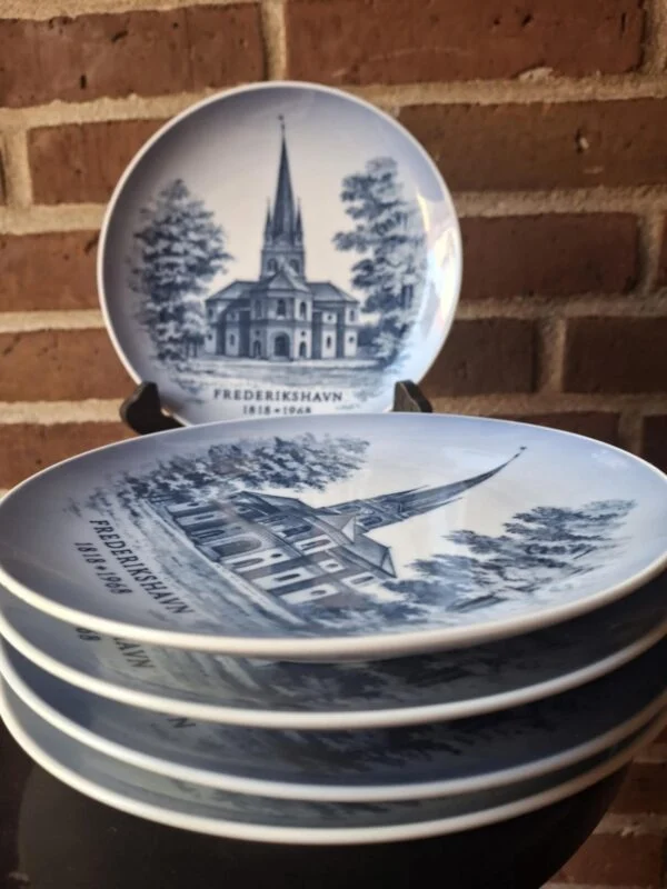 Flere Royal Copenhagen-platter med Frederikshavn Kirke er stablet foran en opretstående platte; varen sælges enkeltvis.