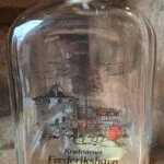 Nærbillede af klar glasflaske med ridser, korkprop og et sort-rødt motiv med teksten Krudttårnet Frederikshavn.