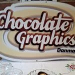 Lyst og brunt Chocolate Graphics Danmark-skilt på en blomsterdug, med glas og chokolade i forgrunden.