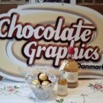 To glas på fod, en skål med chokolade og to flasker Julemjød på en broderet dug foran et Chocolate Graphics Danmark-skilt.