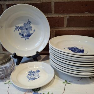 Stor dyb tallerken i hvid porcelæn med håndmalet blåt blomstermotiv i midten. Står på sort holder foran murstensvæg og hvid dug.