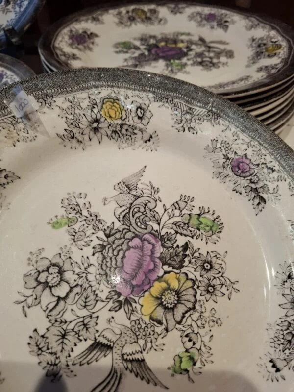 Dyb tallerken med fugle og farvede blomster set skråt ovenfra; flere beslægtede tallerkener står bagved.